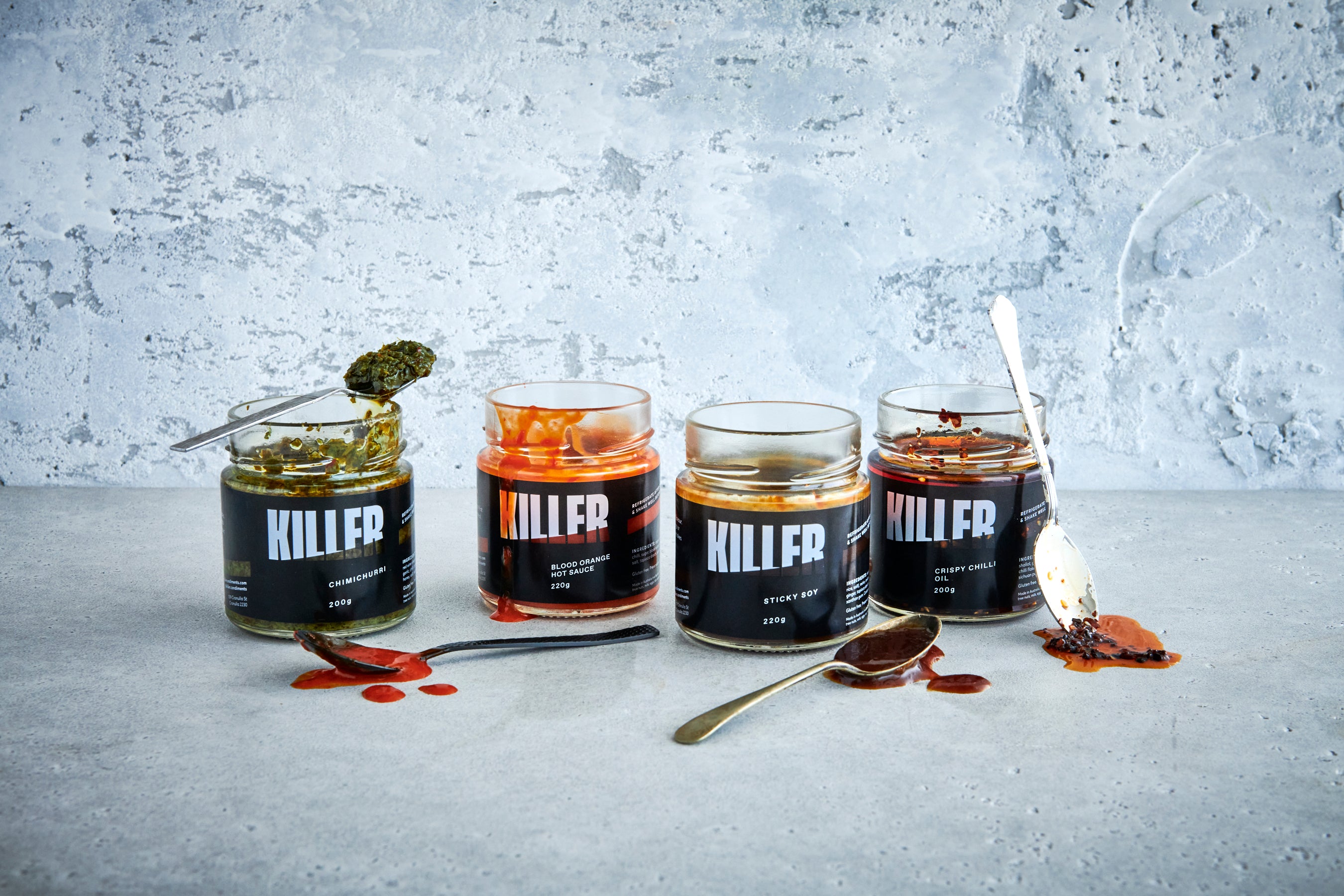 Captivating lifestyle shot of the full range of Killer Condiments artisanal sauces – open jars of Chimichurri, Blood Orange Hot Sauce, Sticky Soy, and Crispy Chilli Oil – tantalizingly dripping down the sides, with spoons lavishly coated in vibrant sauces, set against a minimalist background to accentuate the rich and alluring textures of these gourmet delights.